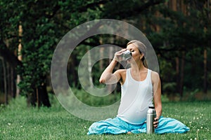 Pregnant yoga in the lotus position with thermos drinking tea. in the park on the grass , outdoor, health woman, female.