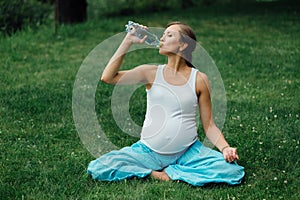Pregnant yoga woman drinking water from a bottle, in the lotus position. park ,grass ,.outdoor, forest.