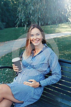 pregnant young Caucasian woman sitting on bench in park outside and drinking coffee from paper cup