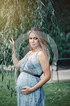 Pregnant young Caucasian woman wearing long blue dress posing in park outside.