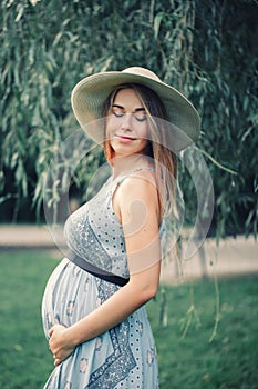 Pregnant young Caucasian woman wearing long blue dress and rustic country hat in park outside.