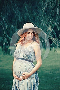Pregnant young Caucasian woman wearing long dress and rustic country hat in park outside.