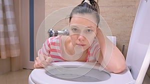 Pregnant young tired woman is vomiting in toilet sitting on the floor at home and looking at camera.