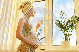 Pregnant young woman with bath towel on head with cup of tea looking out window