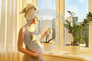 Pregnant young woman with bath towel on head with cup of tea looking out window