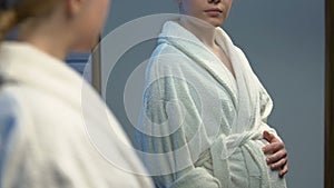Pregnant young woman in bathrobe looking at tummy in mirror, baby expectation