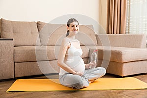 Pregnant young woman with bottle of water while sitting in lotus position on yoga mat. Maintaining water balance at coronavirus