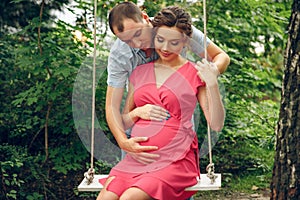 A pregnant young woman and her husband. A happy family sitting on a swing, holding belly. pregnant woman relaxing in the park.