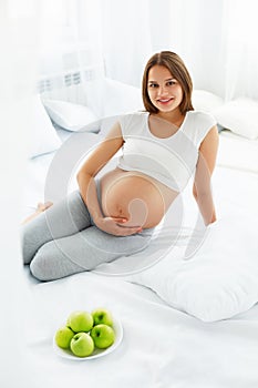 Pregnant Young Woman holding Apple while sitting on the Bed. Hea