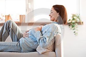 Pregnant Young Woman Hugging Belly Resting On Sofa At Home