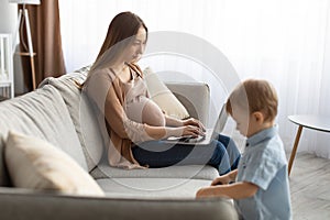 Pregnant young woman working on laptop computer while spending time with her little son at home