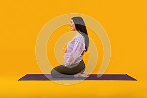 Prenatal yoga. Pregnant lady sitting on fitness mat and touching belly, yellow studio background, side view