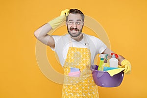 Preoccupied man househusband in apron rubber gloves hold basin with detergent bottles washing cleansers doing housework