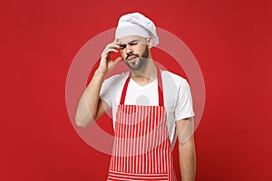 Preoccupied tired young male chef cook or baker man in striped apron toque chefs hat posing isolated on red background