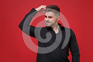Preoccupied young bearded guy 20s in classic black shirt posing isolated on red wall background studio portrait. People