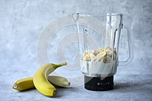 Preparation of a banana cocktail. Glass blender with banana slices and milk on a bluish gray background. Next to the blender are t