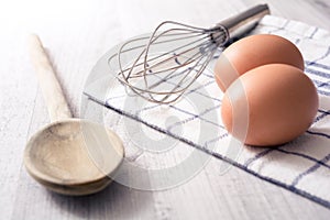 Prepare Breakfast Eggs Concept With 2 Eggs, A Wooden Spoon, An Eggbeater And A Towel On A Kitchen Table