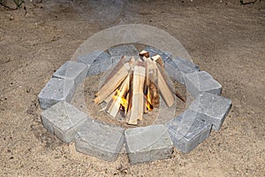 Prepared chopped pieces of wood to make a fire in a stone circle on the sand, visible flames.