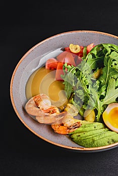 Prepared shrimps or prawns with boiled eggs, avocado, fresh tomatoes and arugula in a plate over black background