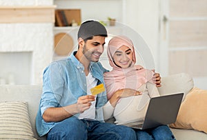 Preparing for childbirth. Loving muslim pregnant couple using laptop computer and credit card, sitting on sofa