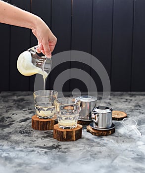 Preparing coffee Vietnamese style. Woman hand pouring condensed milk into glasses on dark background