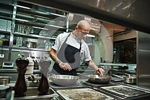 Preparing food. Handsome and confident chef with several tattoos on his arms frying ingredients for his dish in a