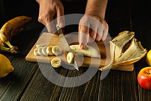 Preparing a fruit dish on the kitchen table. Chef hands using a knife to slice a banana on a wooden cutting board