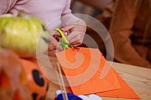 Preparing for Halloween. Close up of a girl's hands cutting out a bat shaped mask on orange paper. Girl at the table