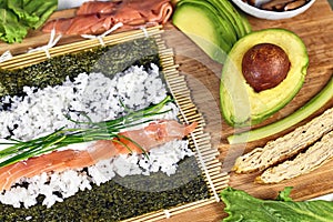 Preparing homemade sushi with Nori algea sheet on bamboo mat and ingredients like smoked salmon, avocado, wild chive