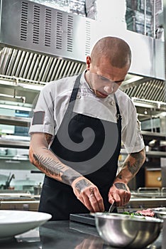 Preparing a salad. Handsome chef with tattoos on his arms cutting cucumber and other food ingredients while standing in