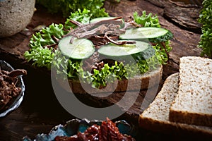 Preparing sandwich with meat, dark bread, fresh green salad, dry tomatoes and fresh cucumbers on rustic wooden table surface