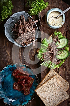 Preparing sandwich with meat, dark bread, fresh green salad, dry tomatoes and fresh cucumbers on rustic wooden table surface