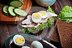 Preparing sandwich with meat, dark bread, fresh green salad, eggs and cucumbers on rustic wooden table surface