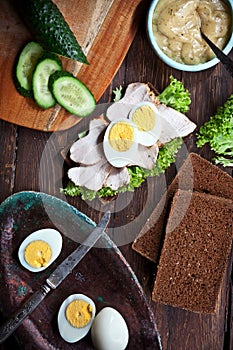 Preparing sandwich with meat, dark bread, fresh green salad, eggs and cucumbers on rustic wooden table surface