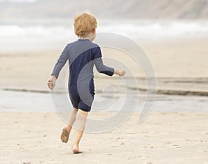 Preschool aged boy in a blue swimming suit running along a sandy shoreline