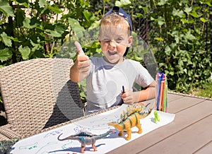 preschool boy draws toy dinosaurs with felt-tip pen while sitting at table in garden