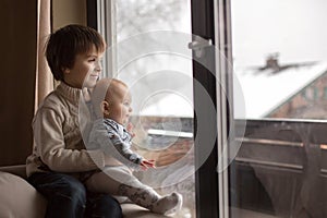 Preschool boy, holding his baby brother, sitting by the window i