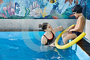 Preschool boy learning to swim in pool with foam noodle with young trainer