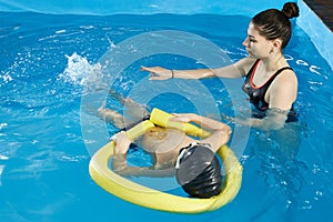 Preschool boy learning to swim in pool with foam noodle with young trainer