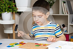 A preschool child plays with a mosaic, builds figures on a board, hammering nails with a hammer on multi-colored parts