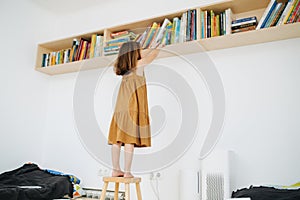 Preschool girl got up on a stool to get a book off the shelf