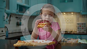 Preschool girl in pink t-shirt eating homemade pizza standing on blue kitchen at home. Kid enjoy eating pizza with hands