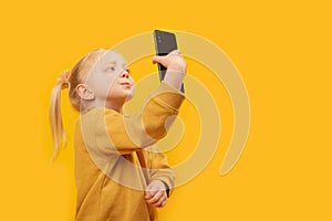 Preschool girl takes selfie on yellow background. New generation and modern tech overuse. Child uses mobile phone