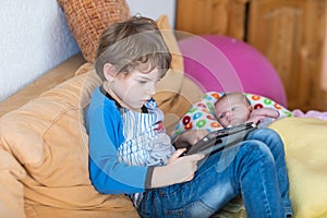 Preschool kid boy playing games tablet computer. Cute newborn baby looking on brother.