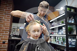 Preschooler boy getting haircut in barbershop. Children hairdresser with professional tools - comb and scissors. Cutting hair for