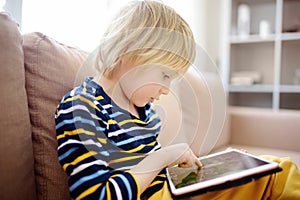 Preschooler boy is learning on distance by tablet or playing pc game sitting on couch. Homeschool education and entertainments for