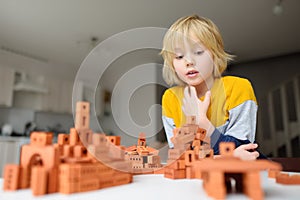 Preschooler boy is playing with real small clay bricks at the table at home. Child having fun and building smart constructions.