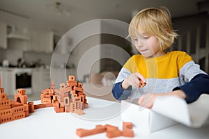 Preschooler boy is playing with real small clay bricks at the table at home. Child having fun and building smart constructions.