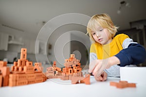 Preschooler boy is playing with real small clay bricks at the table at home. Child having fun and building smart constructions.