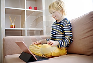 Preschooler boy is watching movie or cartoon movie from tablet sitting on couch. Entertainments for kids while coronavirus